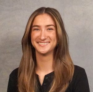 Picture of young woman with light brown medium length hair, green eyes, small nose piercing in black shirt.