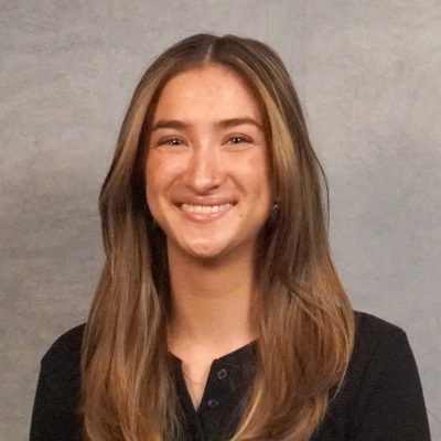 Picture of young woman with light brown medium length hair, green eyes, small nose piercing in black shirt.
